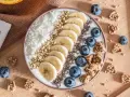 Un bol de smoothie aux bananes, myrtilles, noix de coco râpée et graines de chia, parfait pour un déjeuner au bureau sain et gourmand.