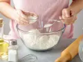 Une personne ajoute une poudre blanche à de la farine dans un bol. Une cuillère et un petit bol sont utilisés. C'est une idée pour par quoi remplacer la levure chimique.