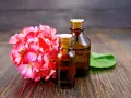 Deux flacons d'huile essentielle sur fond de bois sombre, à côté d'une grappe de fleurs roses. Une solution naturelle contre les moustiques.