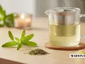 Une théière en verre fumante contient une tisane verveine dorée. Des feuilles fraîches et séchées de verveine, ainsi qu'une tranche de citron, complètent la scène.