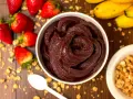 Un bol de sorbet **açaí** onctueux, entouré de fraises, bananes et granola. Une cuillère blanche est posée à côté.