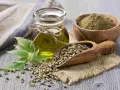 Huile de chanvre cheveux : une fiole d'huile dorée, des graines dans une cuillère en bois, de la poudre verte et une feuille de chanvre sur fond de bois.