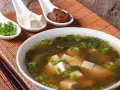 Un bol de soupe miso avec des cubes de tofu, des algues et des oignons verts ciselés. Trois cuillères présentent des ingrédients : oignons verts, tofu et pâte miso.
