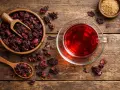 Infusion hibiscus bienfaits : une tasse de thé rouge vif à côté d'un bol de fleurs d'hibiscus séchées et d'une cuillère en bois.