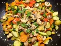 Un wok de légumes asiatique coloré avec des carottes, courgettes, poivrons et champignons, parsemé de graines de sésame et de persil frais.