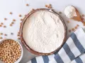 Un bol de pois chiches entiers à côté d'un bol rempli de farine de pois chiche. Une cuillère en bois déborde de cette poudre fine.