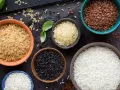 Cinq bols de riz différents : blanc, brun, noir, rouge et riz germé, disposés sur un plateau sombre avec des herbes et des épices.