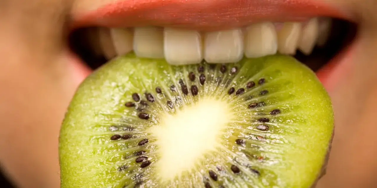 Une tranche de kiwi est mordue par des lèvres roses, ses petites graines noires contrastant avec la chair verte. Une sensation de kiwi langue qui pique.