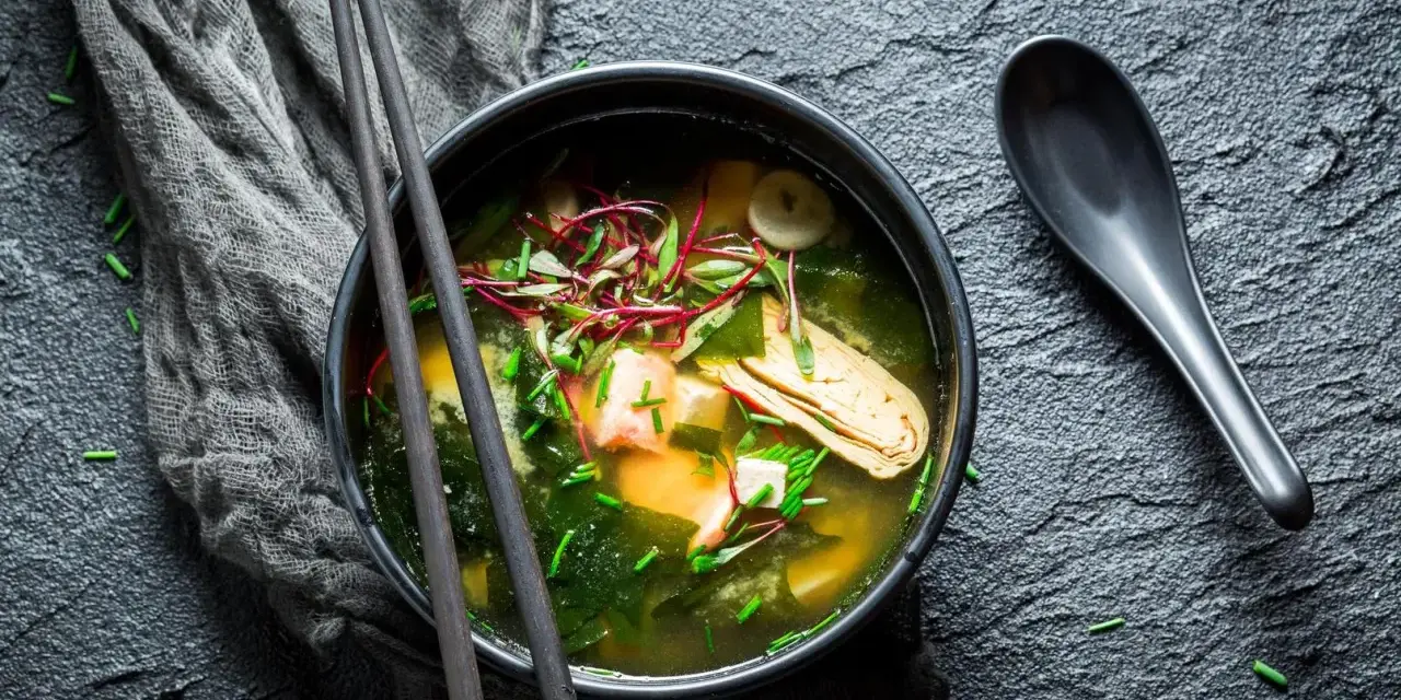 Un bol de bouillon miso fumant, garni de tofu, d'algues et de pousses rouges, prêt à être dégusté avec des baguettes.