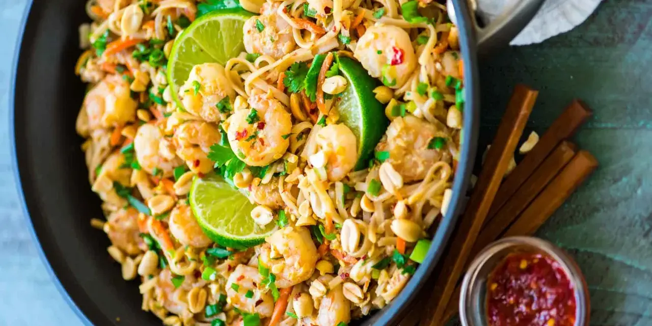 Un plat de recette pad thai crevette appétissant dans un wok, garni de crevettes, de cacahuètes, de coriandre et de quartiers de citron vert.