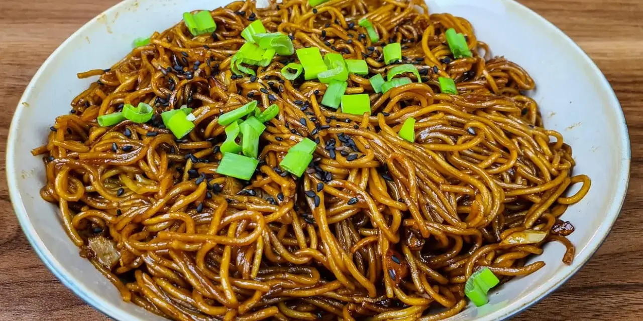 Un plat de nouilles savoureuses, enrobées d'une sauce brune appétissante, garnies de graines de sésame noir et de ciboule fraîche.