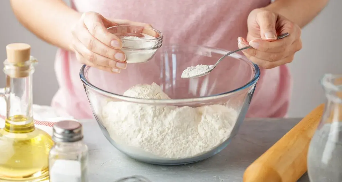 Une personne ajoute une poudre blanche à de la farine dans un bol. Une cuillère et un petit bol sont utilisés. C'est une idée pour par quoi remplacer la levure chimique.