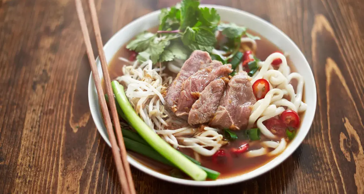 Un bol de soupe nouille de riz avec du bœuf, des germes de soja, des oignons verts et du piment rouge, prêt à être dégusté avec des baguettes.