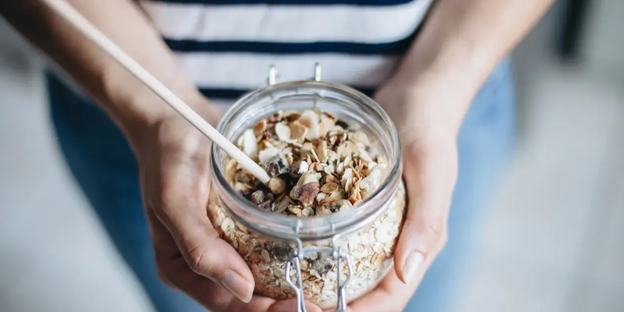 Mains de personne tenant un bocal en verre rempli de muesli maison, avec une cuillère en bois.