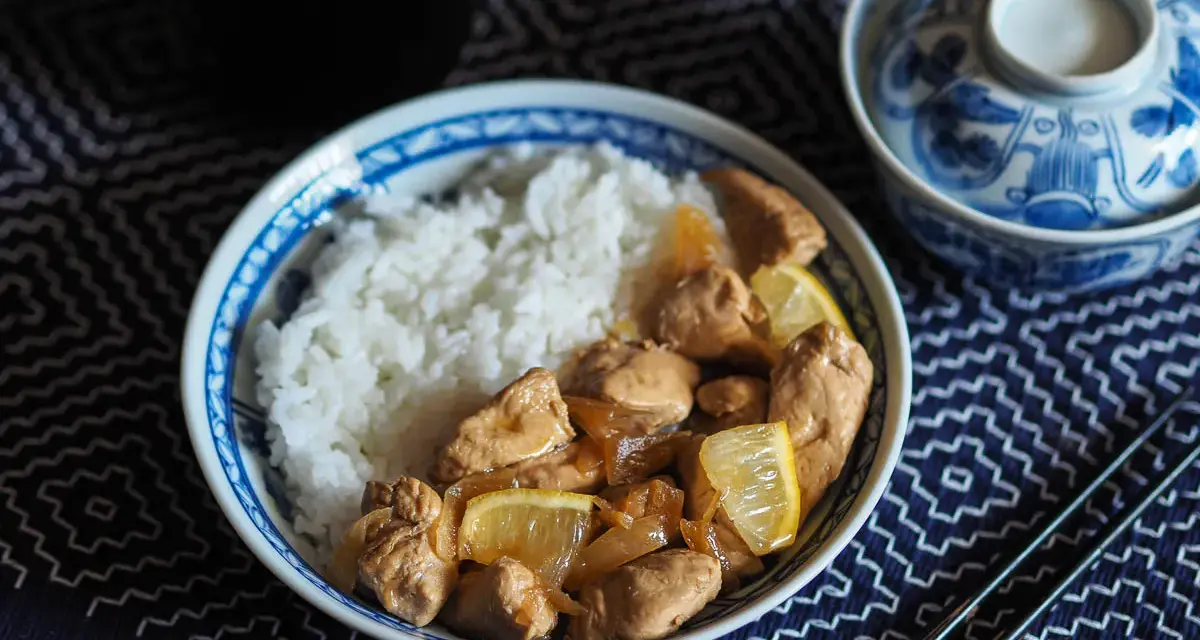 Un plat de riz blanc avec du poulet mariné sauce soja, garni de quartiers de citron. Un bol décoré et des baguettes complètent la scène.
