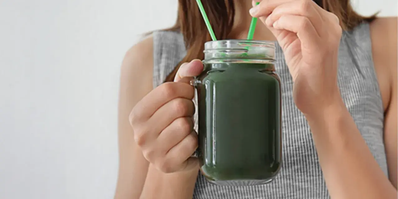 Une femme boit un smoothie vert à la spiruline, un allié pour la santé et la beauté des cheveux.