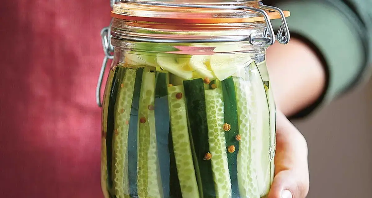 Bocal rempli de concombres coupés en lanières, prêts pour la lacto fermentation. Des grains de moutarde flottent dans la saumure.