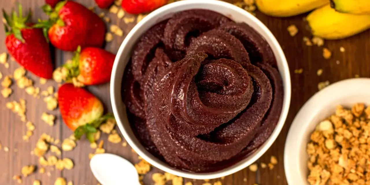 Un bol de sorbet **açaí** onctueux, entouré de fraises, bananes et granola. Une cuillère blanche est posée à côté.