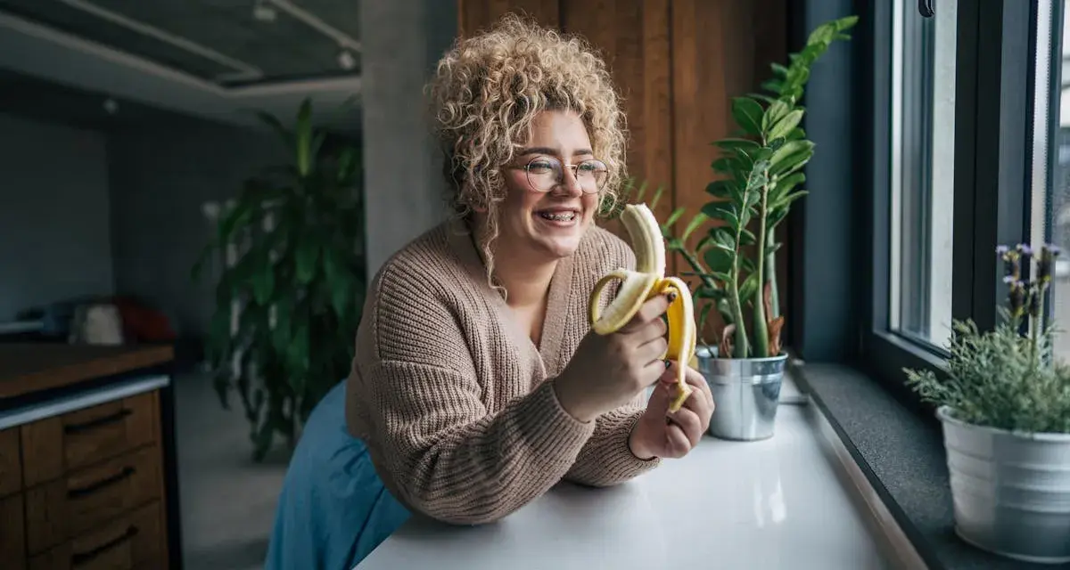 Une femme souriante avec des cheveux bouclés tient une banane, malgré une possible allergie à la banane.