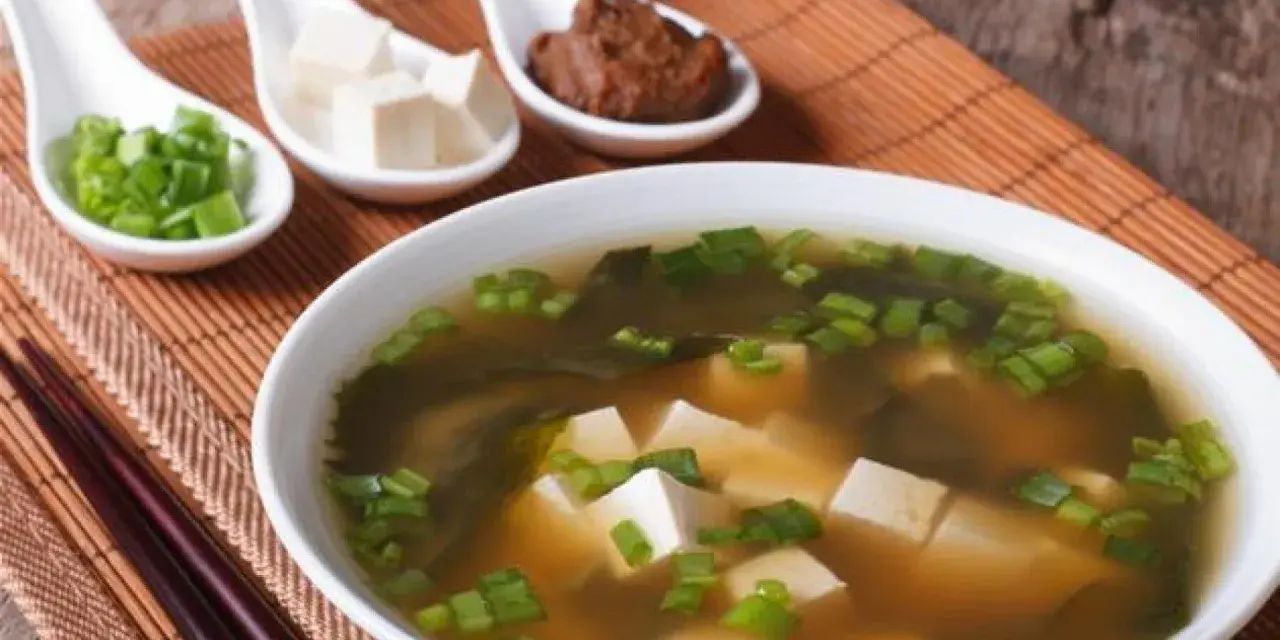 Un bol de soupe miso avec des cubes de tofu, des algues et des oignons verts ciselés. Trois cuillères présentent des ingrédients : oignons verts, tofu et pâte miso.