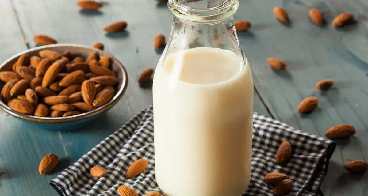 Bouteille de lait végétal d'amande, avec un bol d'amandes et des amandes éparpillées sur une table en bois bleu.