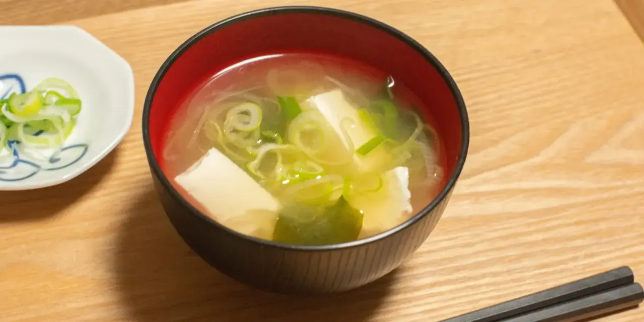 Un bol de soupe japonaise, avec du tofu et des oignons verts, servi avec des baguettes sur un plateau en bois.