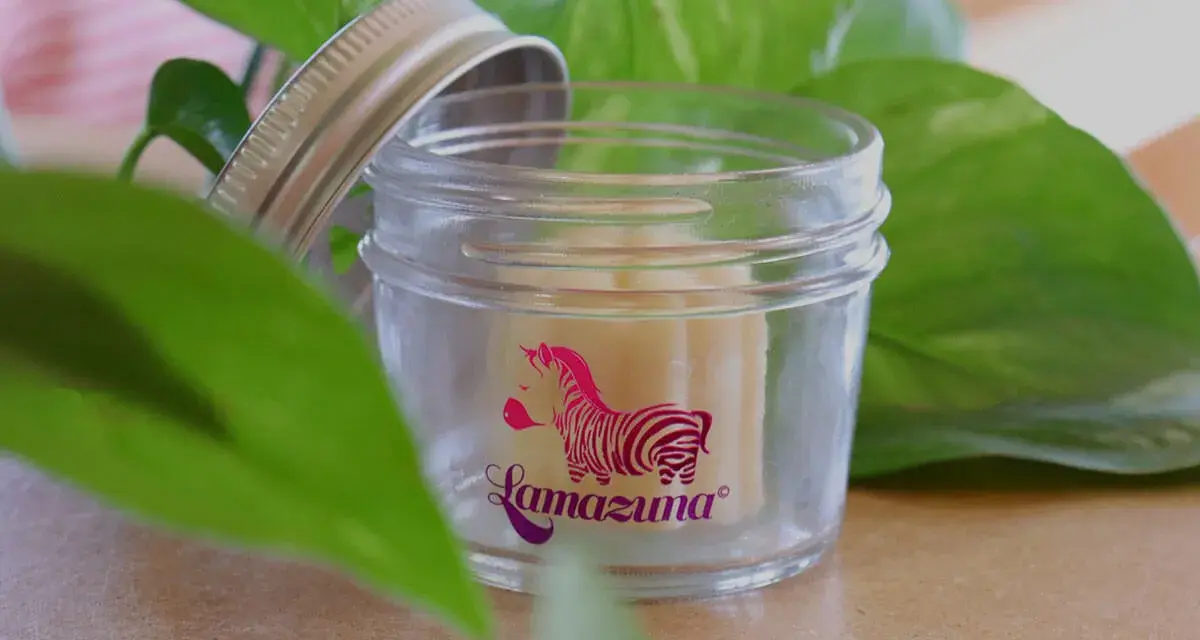 Petit pot en verre ouvert, révélant un baume solide. Le logo Lamazuna, un zèbre rose, orne le verre. Un soin naturel à base de beurre de cacao.