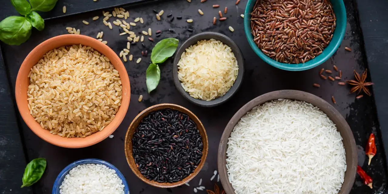 Cinq bols de riz différents : blanc, brun, noir, rouge et riz germé, disposés sur un plateau sombre avec des herbes et des épices.