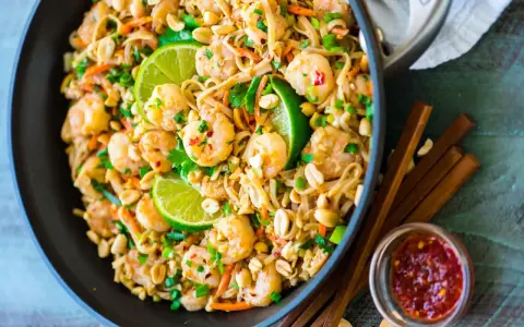 Un plat de recette pad thai crevette appétissant dans un wok, garni de crevettes, de cacahuètes, de coriandre et de quartiers de citron vert.