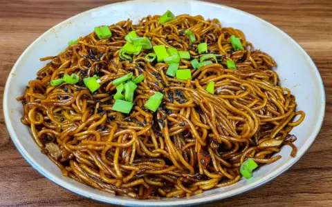 Un plat de nouilles savoureuses, enrobées d'une sauce brune appétissante, garnies de graines de sésame noir et de ciboule fraîche.