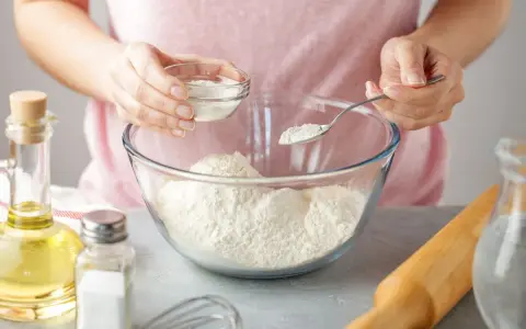 Une personne ajoute une poudre blanche à de la farine dans un bol. Une cuillère et un petit bol sont utilisés. C'est une idée pour par quoi remplacer la levure chimique.