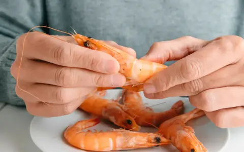 Des mains épluchent une crevette crue, posée sur une assiette avec d'autres crevettes. Peut on manger des crevettes crues ?