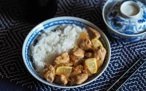 Un plat de riz blanc avec du poulet mariné sauce soja, garni de quartiers de citron. Un bol décoré et des baguettes complètent la scène.