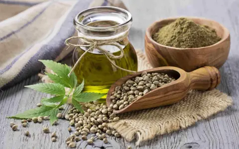 Huile de chanvre cheveux : une fiole d'huile dorée, des graines dans une cuillère en bois, de la poudre verte et une feuille de chanvre sur fond de bois.