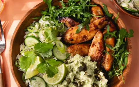 Un bol de riz au miso avec poulet glacé, concombre, coriandre, roquette et une purée verte parsemée de graines.