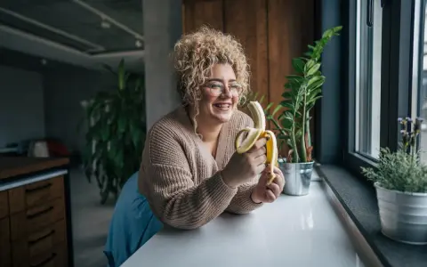 Une femme souriante avec des cheveux bouclés tient une banane, malgré une possible allergie à la banane.
