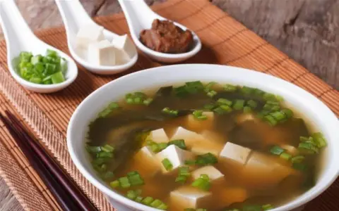 Un bol de soupe miso avec des cubes de tofu, des algues et des oignons verts ciselés. Trois cuillères présentent des ingrédients : oignons verts, tofu et pâte miso.