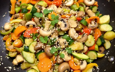 Un wok de légumes asiatique coloré avec des carottes, courgettes, poivrons et champignons, parsemé de graines de sésame et de persil frais.
