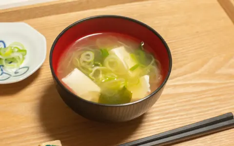 Un bol de soupe japonaise, avec du tofu et des oignons verts, servi avec des baguettes sur un plateau en bois.