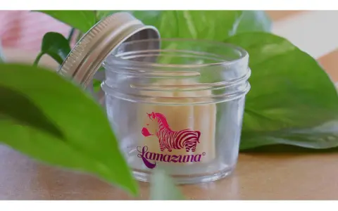 Petit pot en verre ouvert, révélant un baume solide. Le logo Lamazuna, un zèbre rose, orne le verre. Un soin naturel à base de beurre de cacao.