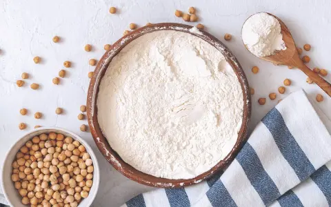 Un bol de pois chiches entiers à côté d'un bol rempli de farine de pois chiche. Une cuillère en bois déborde de cette poudre fine.