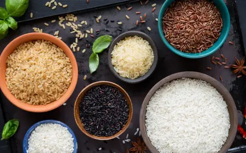 Cinq bols de riz différents : blanc, brun, noir, rouge et riz germé, disposés sur un plateau sombre avec des herbes et des épices.