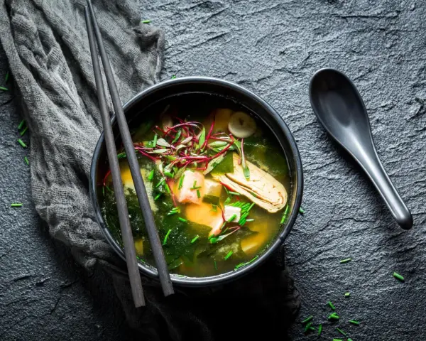 Un bol de bouillon miso fumant, garni de tofu, d'algues et de pousses rouges, prêt à être dégusté avec des baguettes.