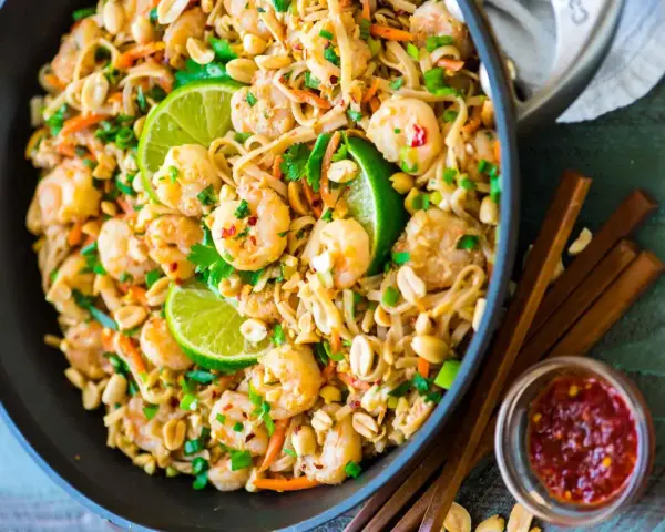Un plat de recette pad thai crevette appétissant dans un wok, garni de crevettes, de cacahuètes, de coriandre et de quartiers de citron vert.