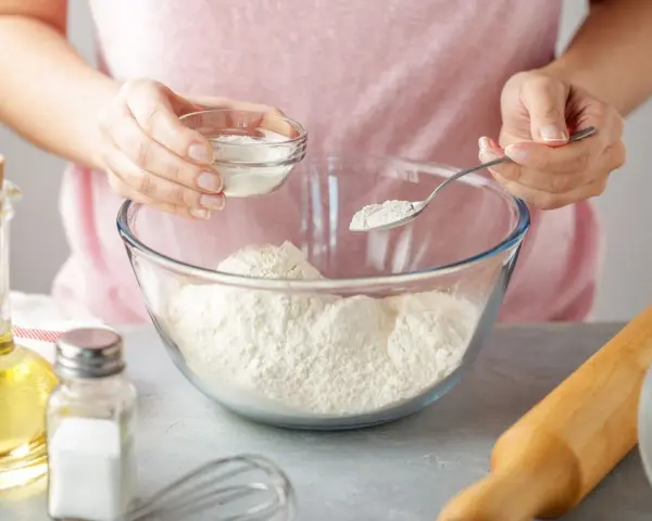 Une personne ajoute une poudre blanche à de la farine dans un bol. Une cuillère et un petit bol sont utilisés. C'est une idée pour par quoi remplacer la levure chimique.