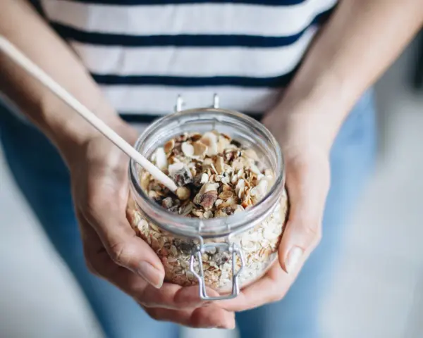 Mains de personne tenant un bocal en verre rempli de muesli maison, avec une cuillère en bois.