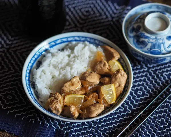 Un plat de riz blanc avec du poulet mariné sauce soja, garni de quartiers de citron. Un bol décoré et des baguettes complètent la scène.