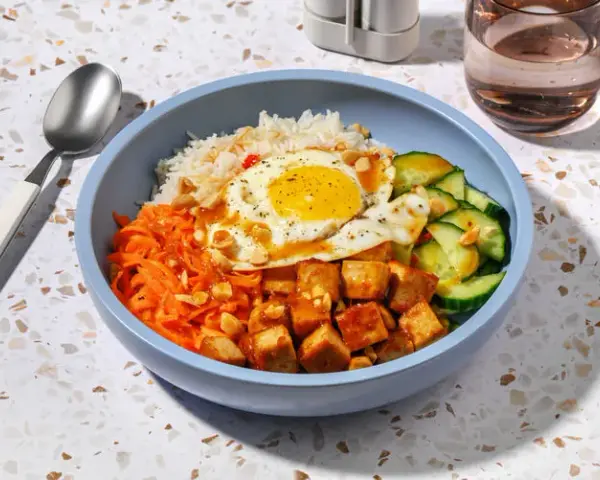 Un bol de bibimbap tofu avec riz, carottes râpées, concombre, œuf au plat et cacahuètes concassées.