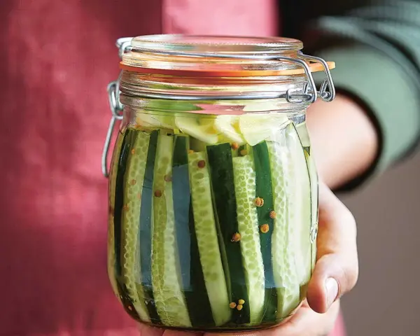 Bocal rempli de concombres coupés en lanières, prêts pour la lacto fermentation. Des grains de moutarde flottent dans la saumure.