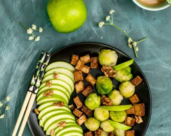 Salade colorée avec des tranches de pomme verte, des choux de Bruxelles, des dés de tofu croustillant et des noix, le tout arrosé d'une vinaigrette miso.